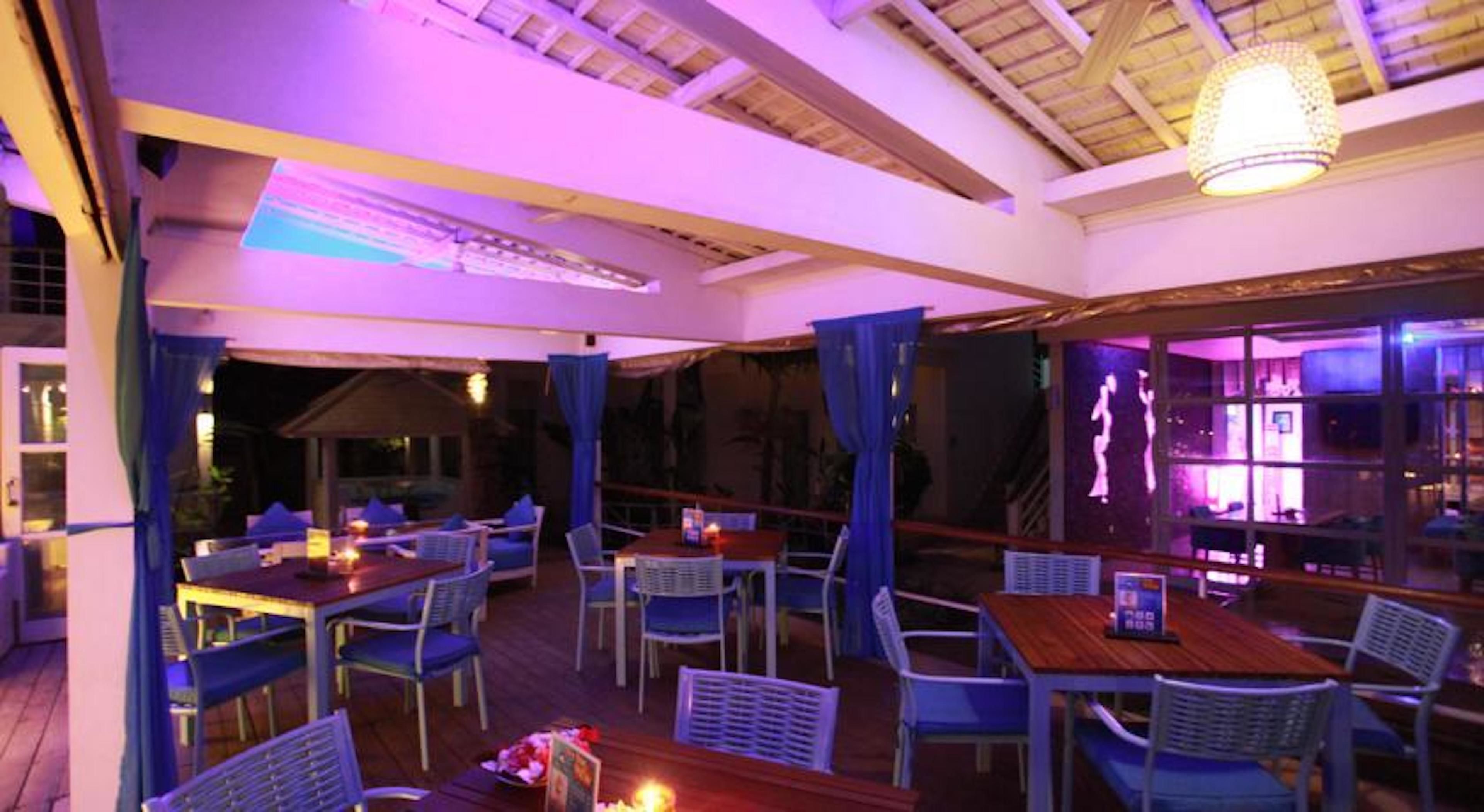 An outdoor dining area with tables and chairs, covered by a roof, and illuminated by ambient lighting.