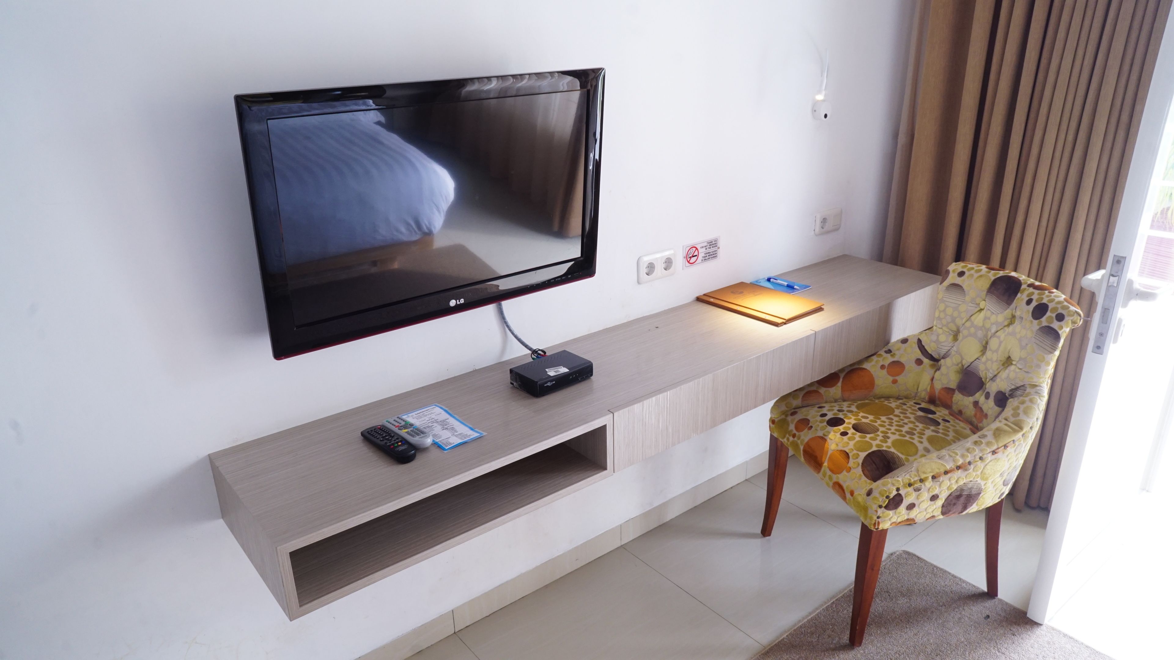 A flat-screen TV mounted on a wall, a remote control, a power strip, and a patterned chair.