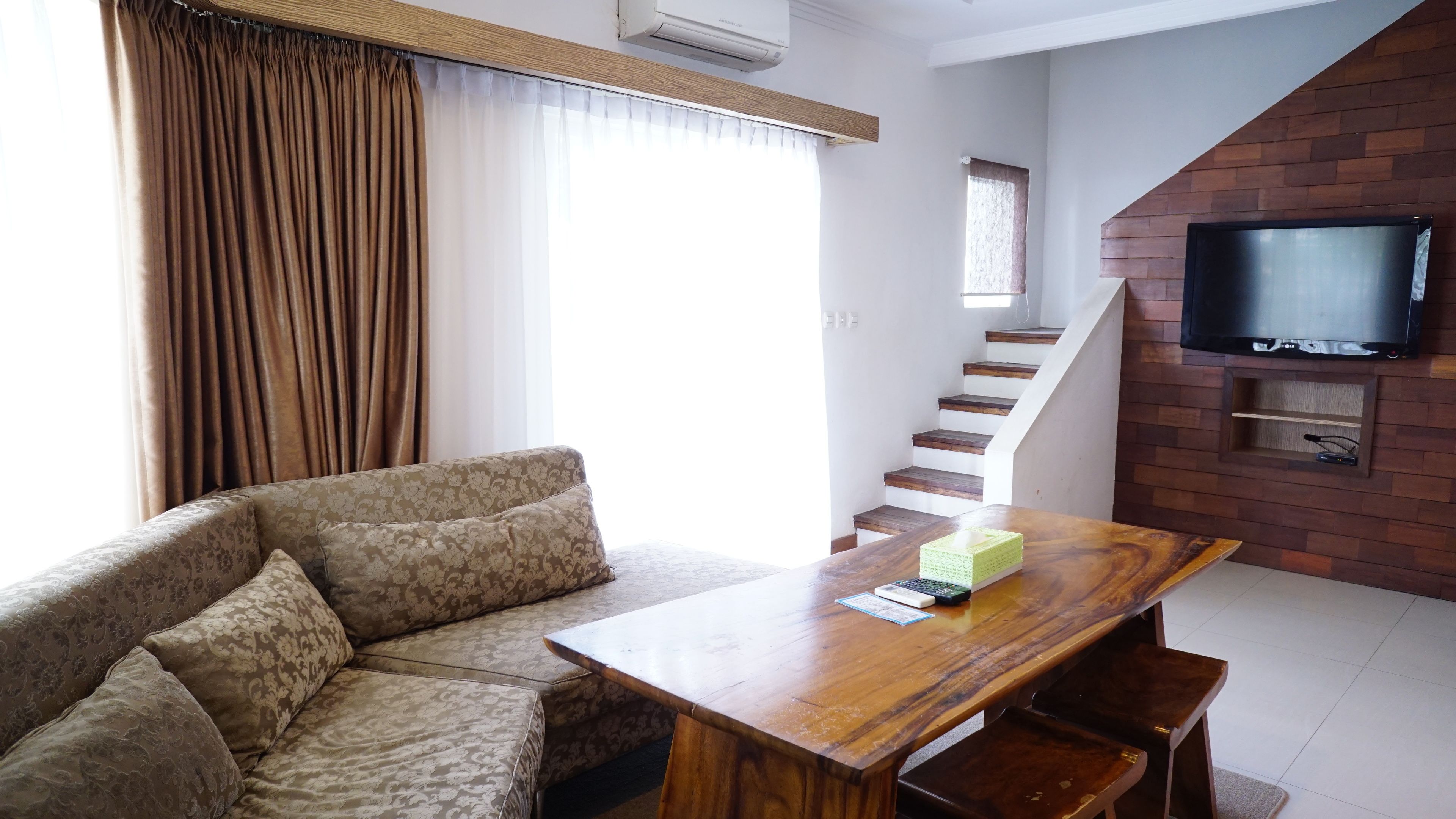 A living room with a sofa, a wooden table, a TV mounted on a brick wall, and a staircase.