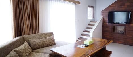 A living room with a sofa, a wooden table, a TV mounted on a brick wall, and a staircase.