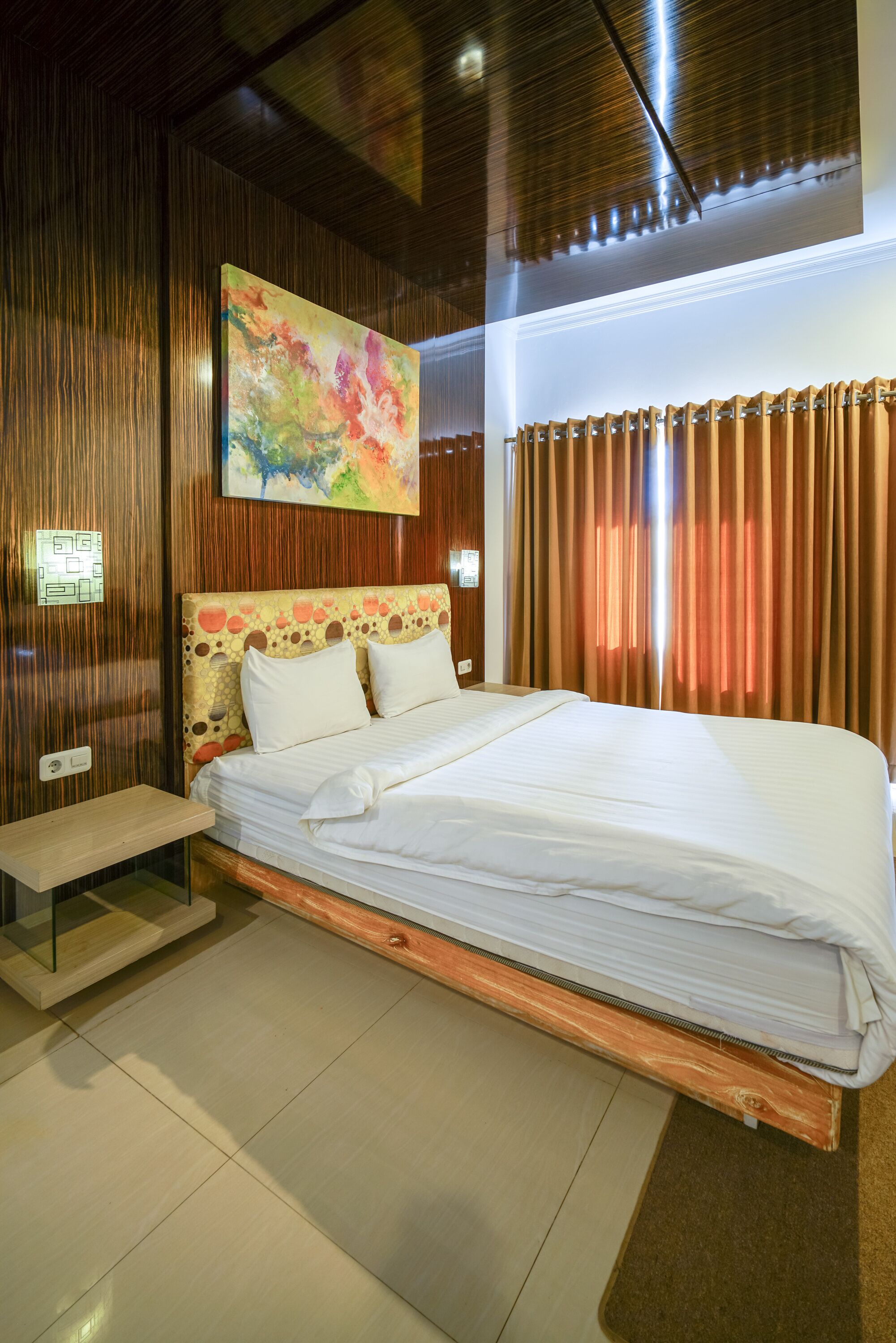 A modern hotel room with a wooden headboard, a large bed, a bedside table, and a colorful abstract painting on the wall.