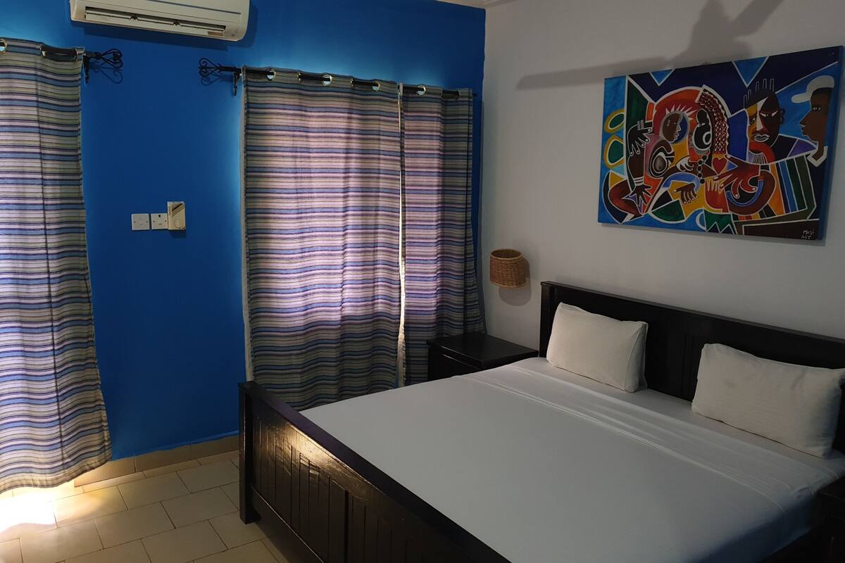 Une chambre à coucher comprenant un lit, des rideaux, un climatiseur, une œuvre d’art accrochée au mur et une table de chevet.