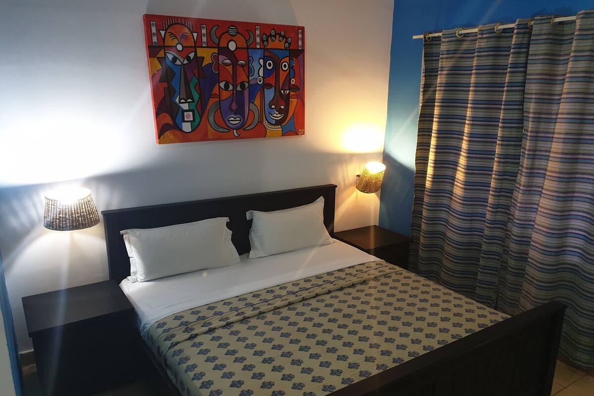Une chambre d’hôtel avec un lit, deux tables de chevet, une œuvre d’art murale et un rideau à rayures.