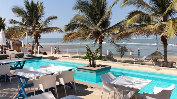 Un espace aménagé au bord de la piscine, avec des chaises longues et une table, offrant une vue sur une plage bordée de palmiers et sur l’océan.