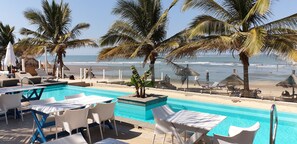 Un área junto a la piscina con sillones de descanso y una mesa, con vista a una playa con palmeras y al océano.