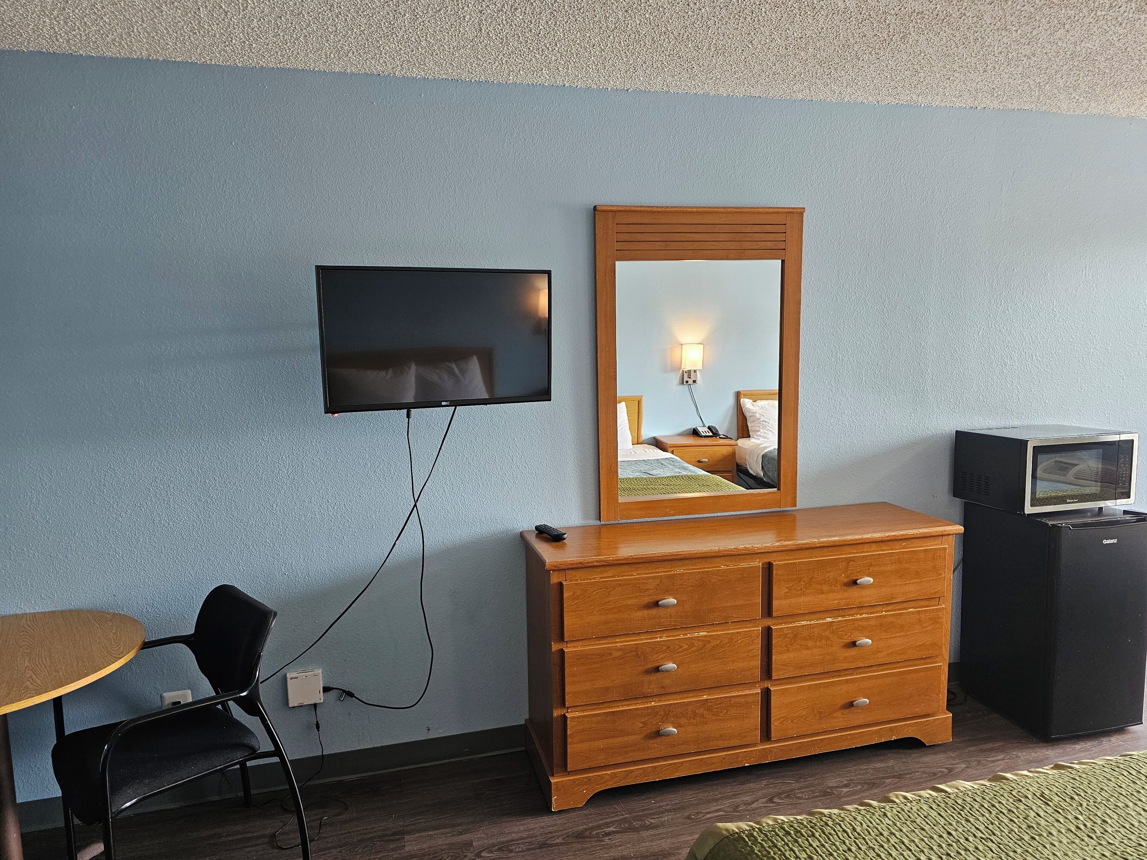 Une chambre d’hôtel équipée d’un téléviseur fixé au mur, d’une commode en bois, d’un four à micro-ondes, d’une chaise et d’une petite table.