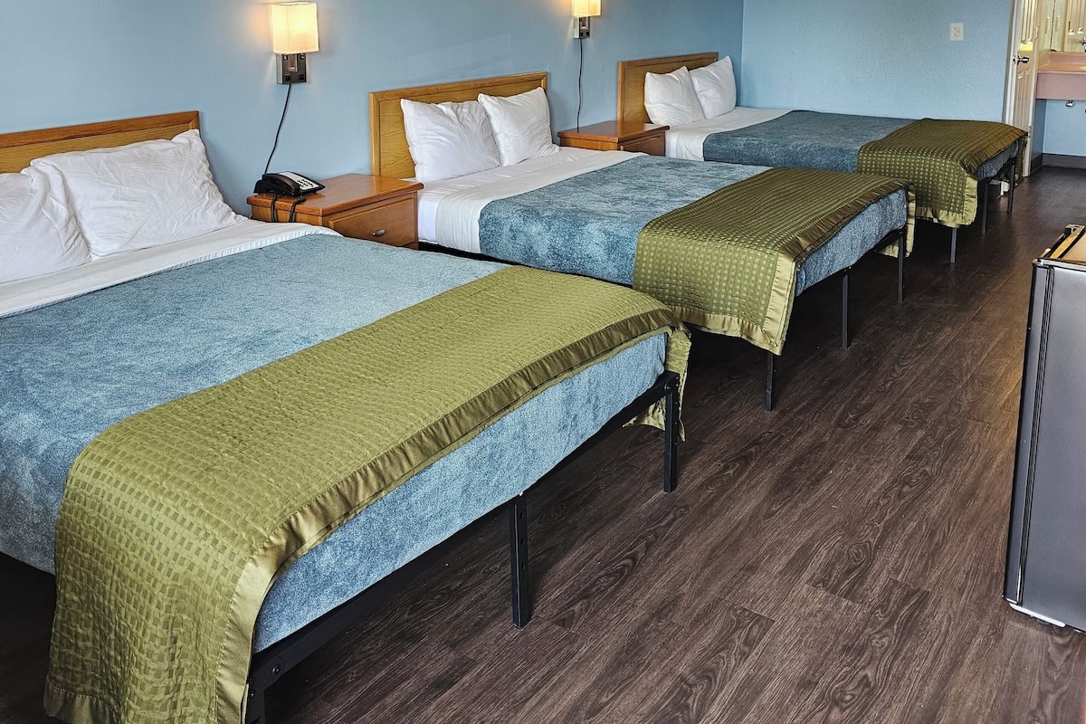 Une chambre d’hôtel avec trois lits, chacun doté d’une tête de lit en bois et de linge de lit blanc. Deux lampes murales éclairent la pièce. La chambre a un plancher en bois et des murs bleu clair.