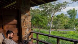 Un homme est assis sur une chaise berçante, sur une terrasse en bois, d’où l’on peut admirer un paysage verdoyant.