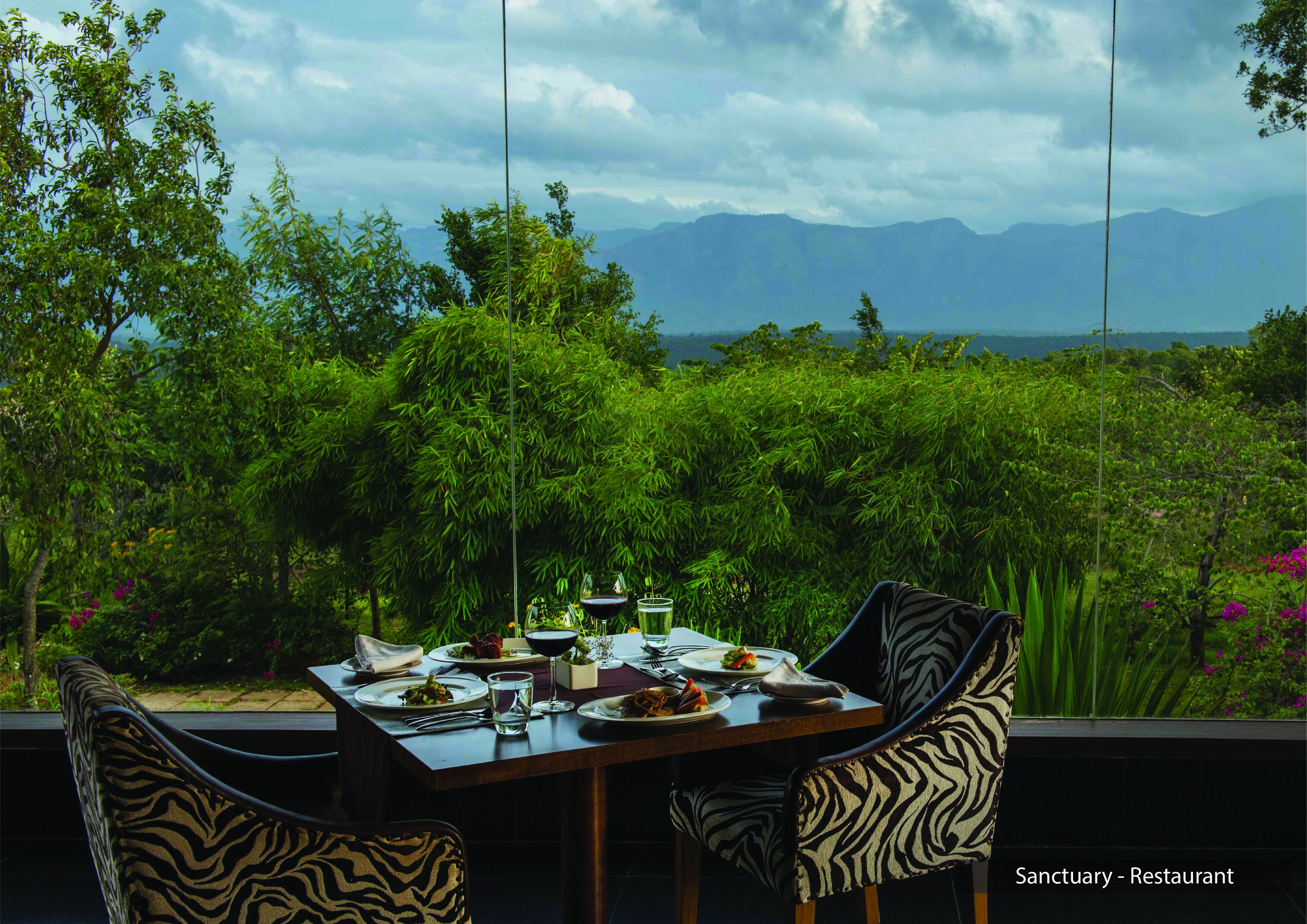 A dining table set with plates, glasses, and cutlery, surrounded by chairs with zebra print upholstery, overlooking a lush garden and distant mountains.