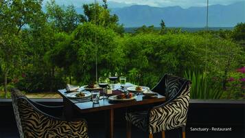 A dining table set with plates, glasses, and cutlery, surrounded by chairs with zebra print upholstery, overlooking a lush garden and distant mountains.