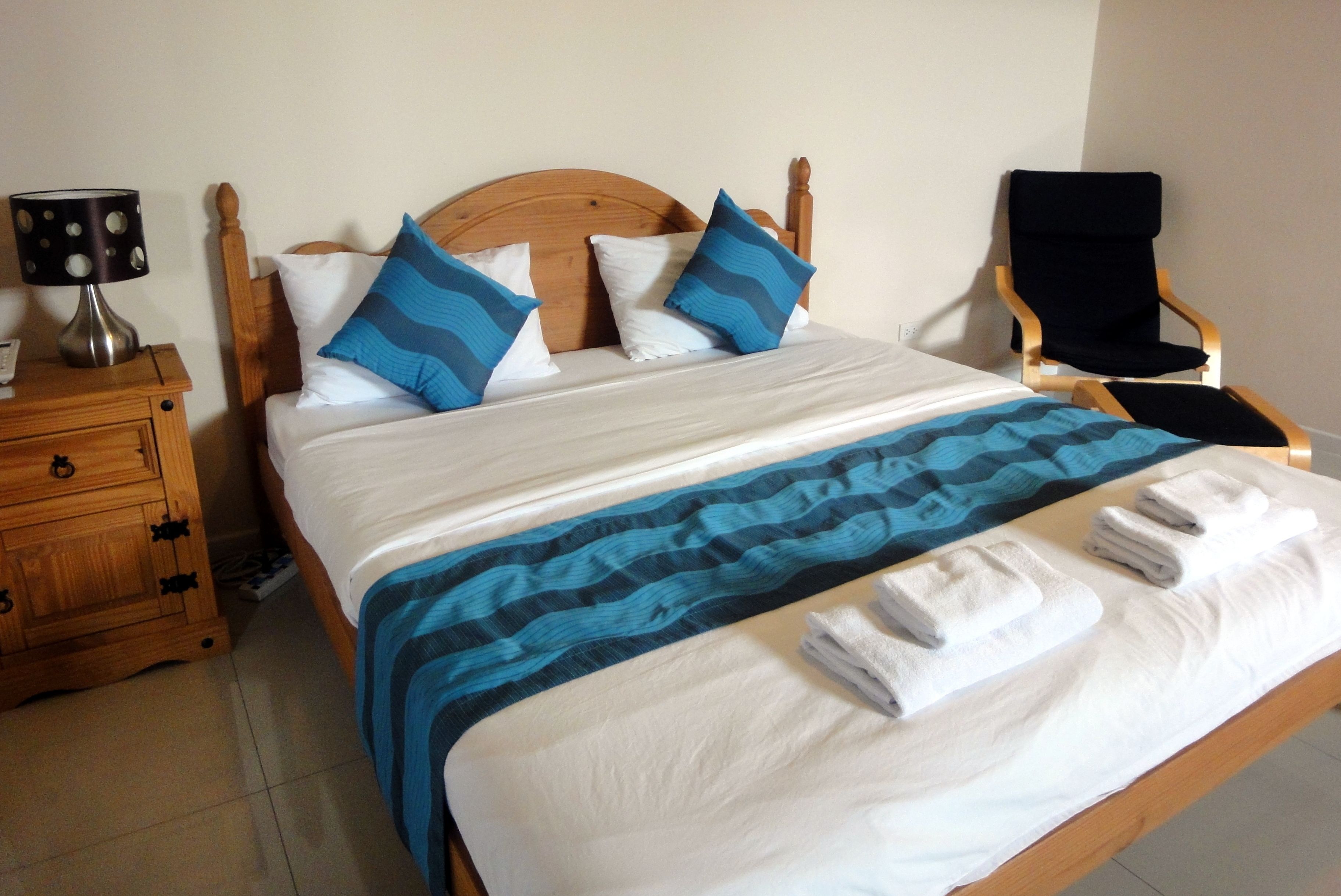 Una cama bien hecha con una manta azul y blanca a rayas, dos almohadas con diseños azules y blancos, una mesita de noche de madera con una lámpara y una silla.