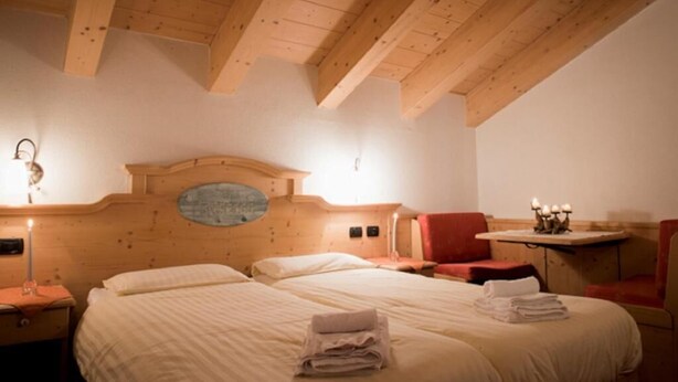 Quarto de hotel acolhedor com teto de madeira, cama grande com toalhas dobradas, mesa pequena com velas e uma cadeira vermelha.