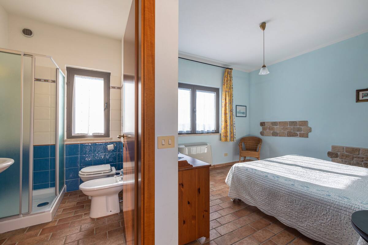 Een badkamer met douche, toilet en wastafel, en een slaapkamer met bed, dressoir en raam.