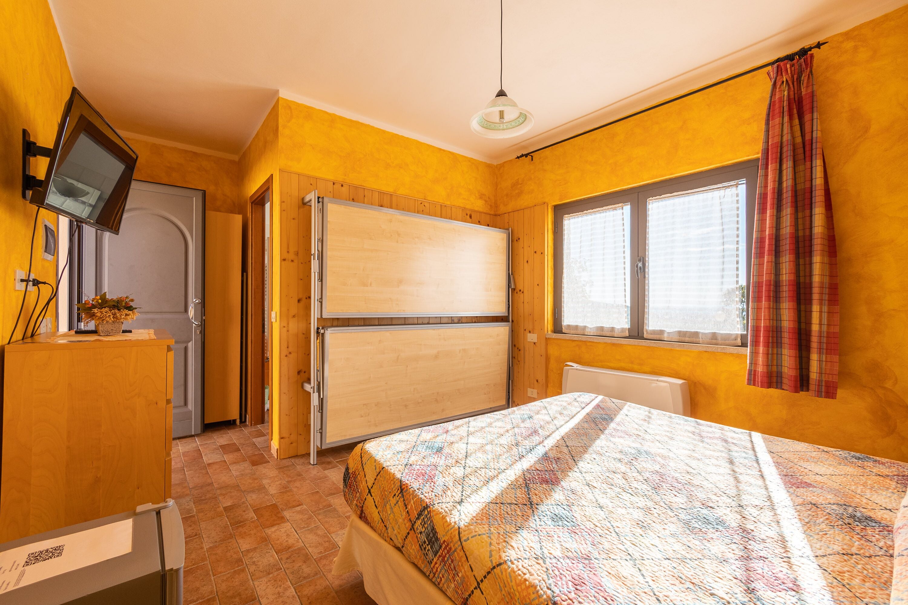 Une chambre à coucher avec un lit, un téléviseur fixé au mur, une fenêtre avec des rideaux et une porte.
