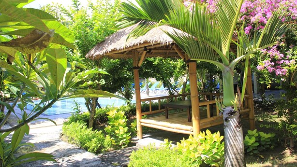 A thatched-roof gazebo with a bench, surrounded by lush greenery and a pool. - Vico Hotel (Nusa Dua)