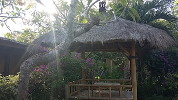 A thatched-roof structure with a wooden bench and a small table, surrounded by lush greenery and purple flowers.