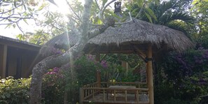 A thatched-roof structure with a wooden bench and a small table, surrounded by lush greenery and purple flowers.