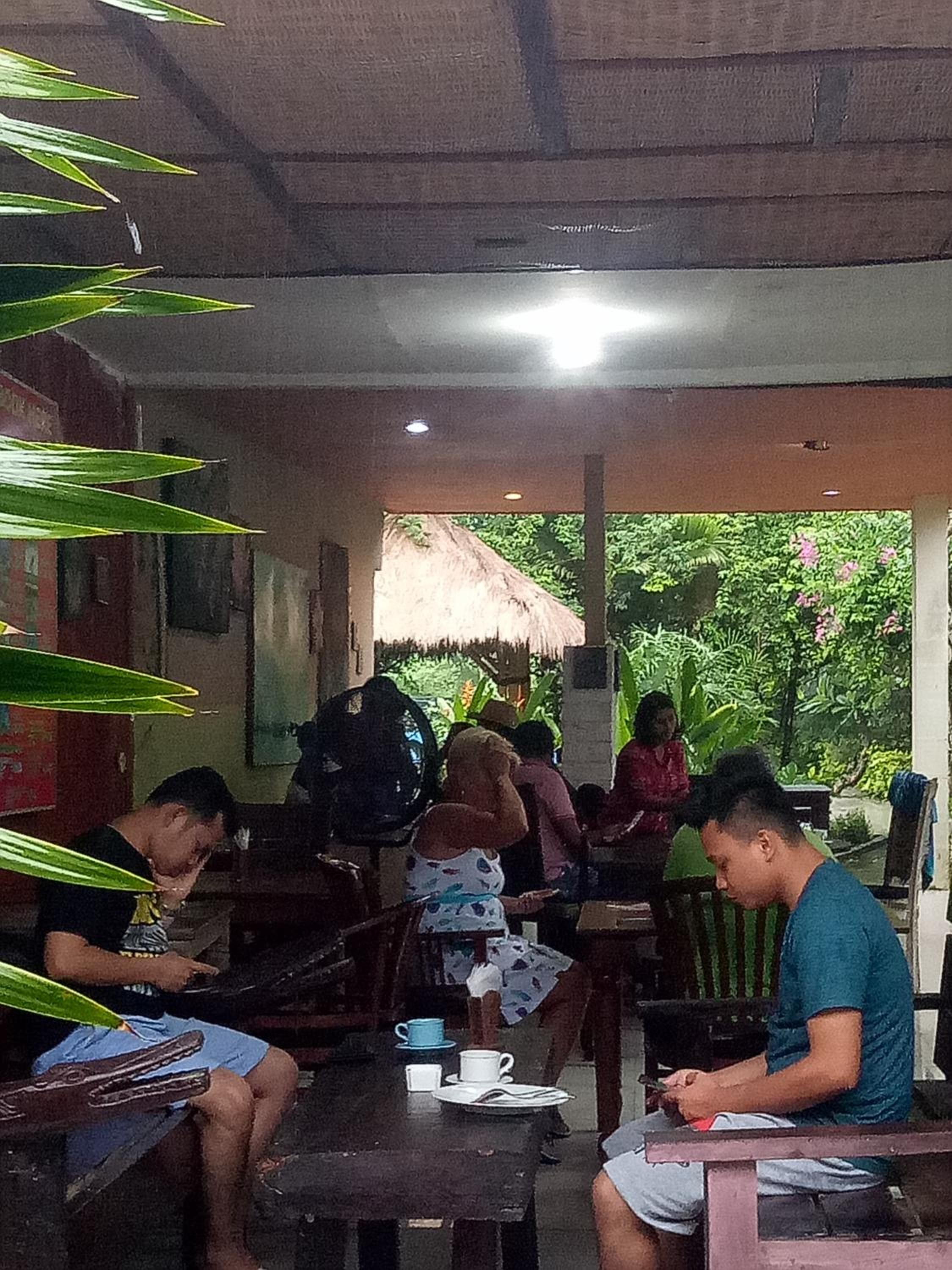 An indoor dining area with tables and chairs, a thatched roof structure, and greenery visible through the windows.