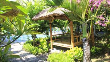 A thatched-roof gazebo with a bench, surrounded by lush greenery and a pool.