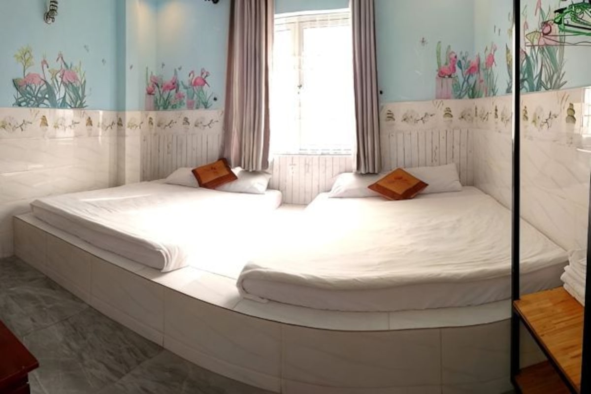 A double bed with white bedding, a wooden nightstand, and a window with curtains.