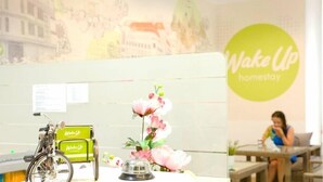 Una recepción con una bicicleta, una campana y un arreglo floral. Una mujer está sentada en una mesa al fondo.