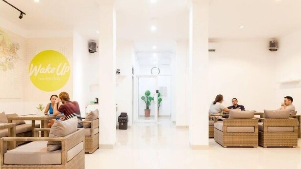 Un moderno vestíbulo de hotel con mobiliario de mimbre, una exhibición floral central y un letrero que dice 'Wake Up'.