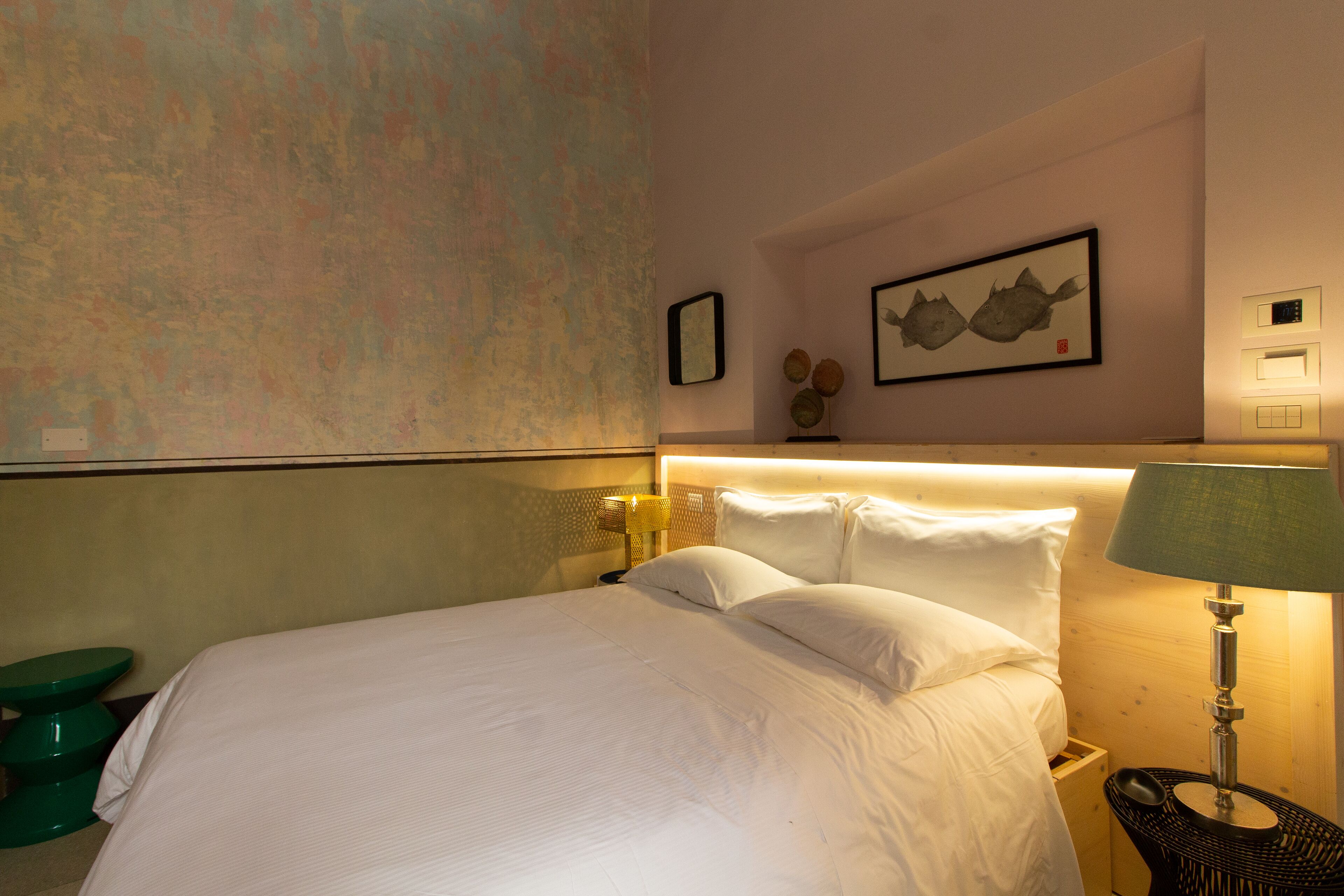 Une chambre d’hôtel avec un lit bien fait, une lampe de chevet, une petite table et une étagère fixée au mur, ornée d’objets décoratifs.