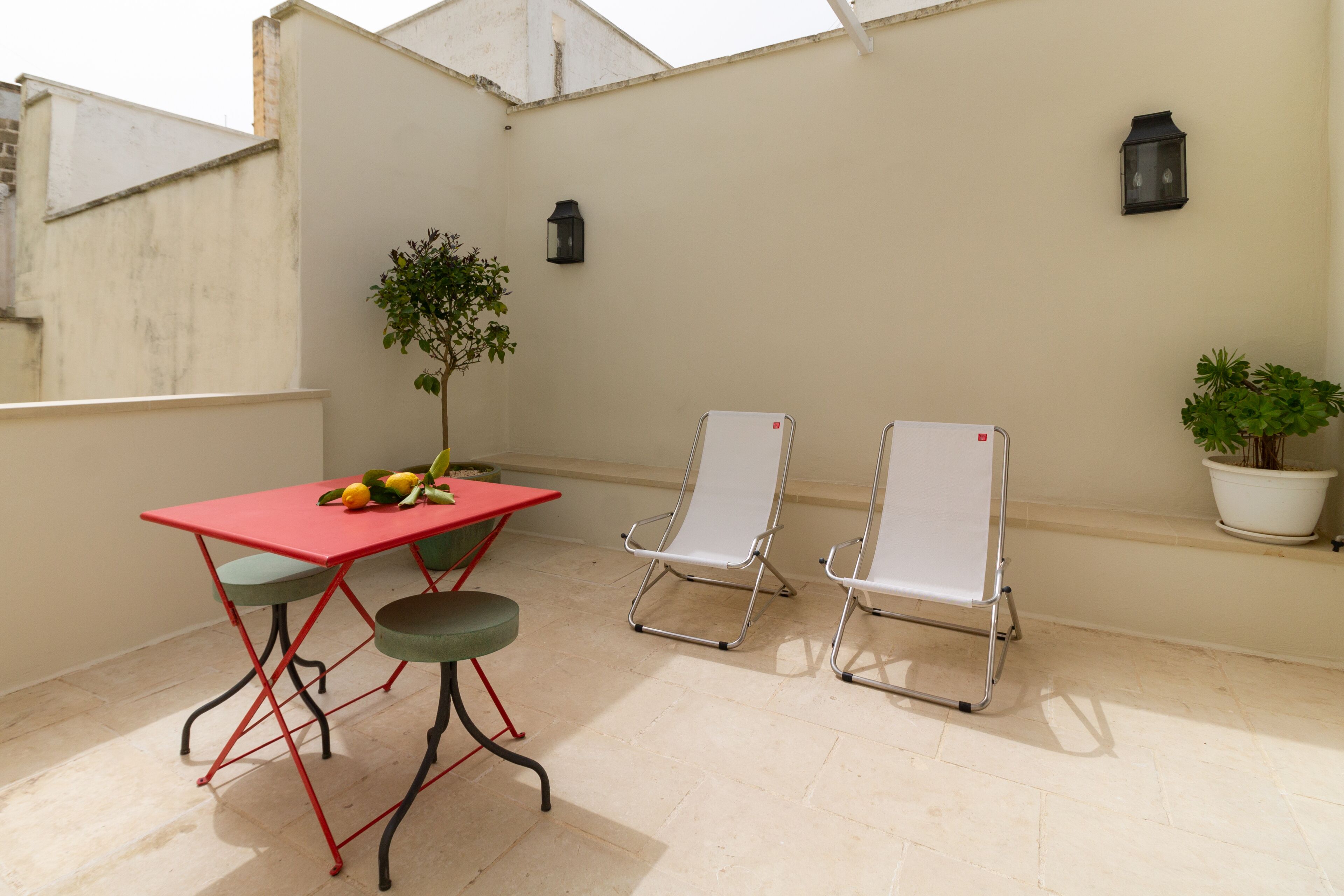 A rooftop terrace with a red table, two white chairs, a potted plant, and two wall-mounted lanterns.