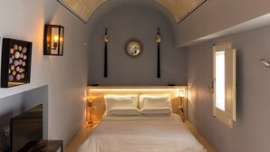 Un dormitorio con una cama, lámparas de noche, un espejo, un televisor montado en la pared y un taburete pequeño rojo.