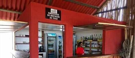 Un área de bar con un letrero que dice "AMBASSADE DE BRETAGNE AU TOGO". Hay taburetes de madera y un refrigerador lleno de bebidas.