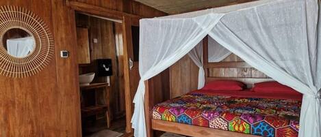 Una habitación de madera con una cama bajo un dosel blanco, una colcha de colores y un espejo con un diseño de sol en la pared.