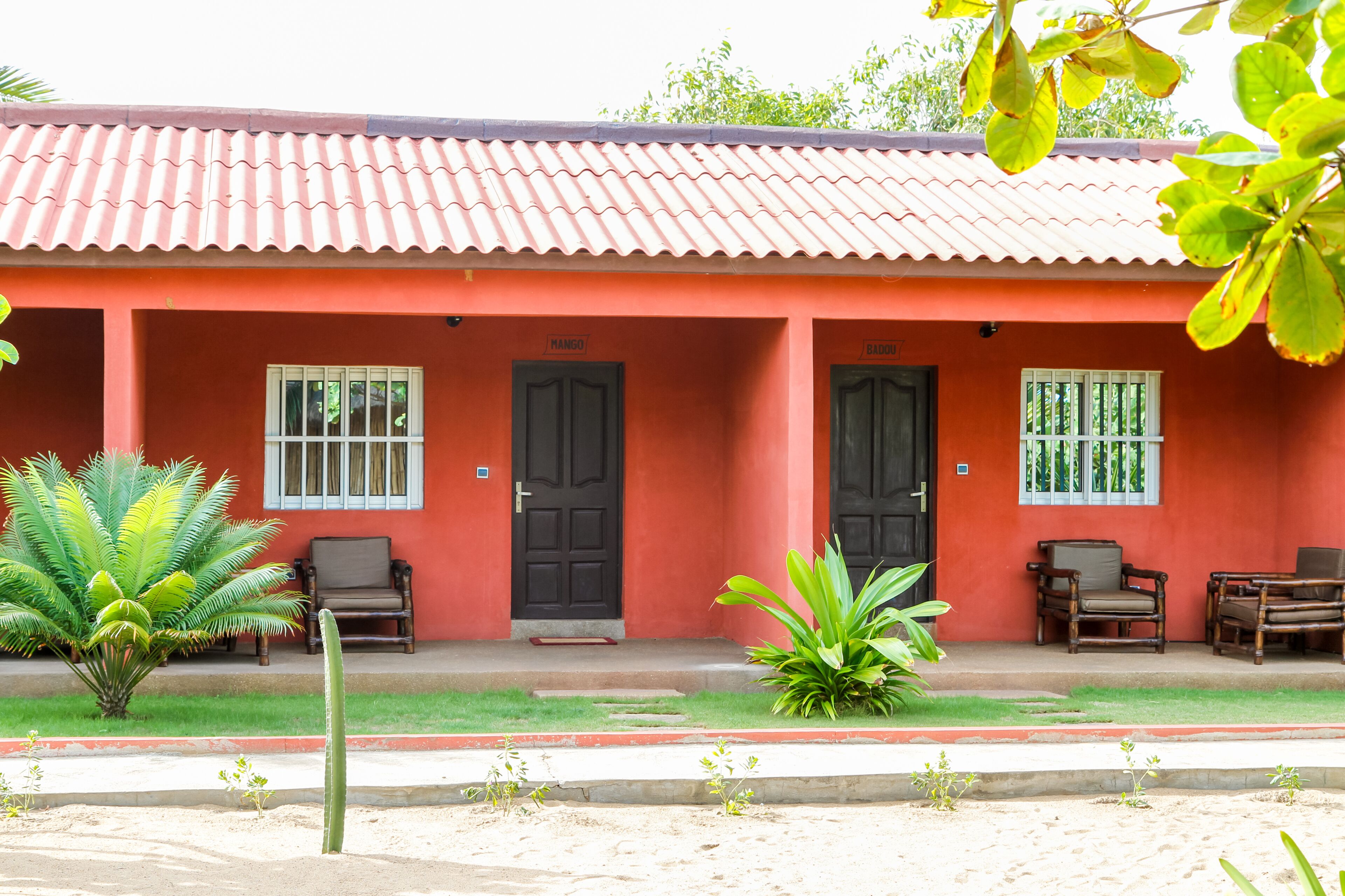 Un bâtiment rouge avec deux portes noires, un petit porche avec des chaises et un cactus au premier plan.