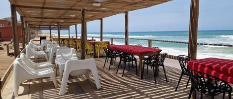A beachside patio with plastic chairs and tables, a wooden deck, and a view of the ocean.