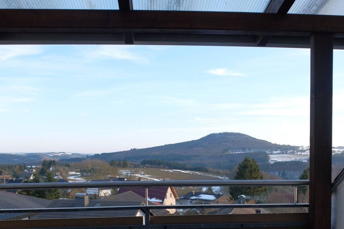 Ein Balkon mit Blick auf einen entfernten Berg, ein Tisch mit einer Flasche und Gläsern sowie ein Korbstuhl.