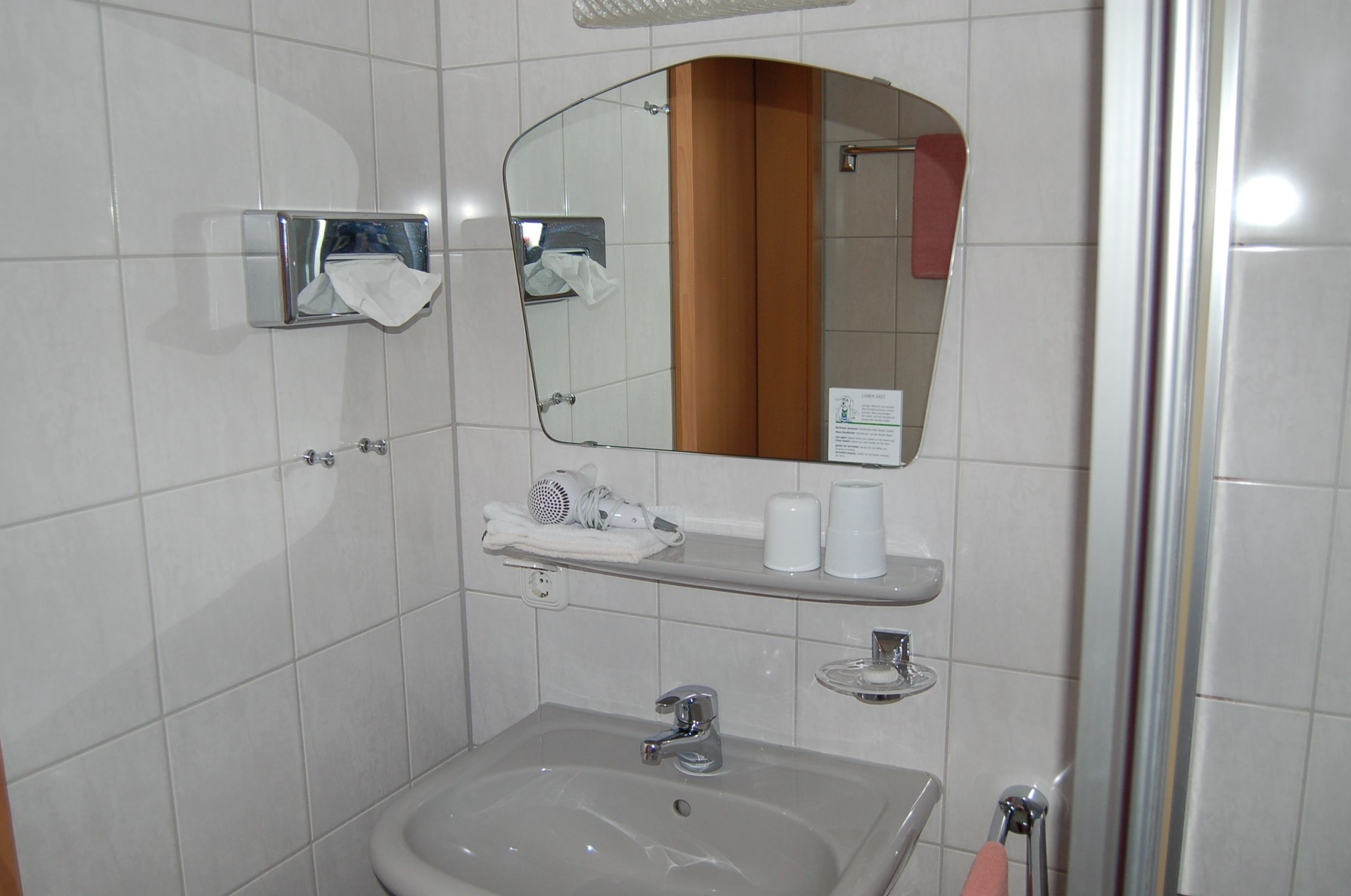 A bathroom with a sink, mirror, towel dispenser, and a shower area.
