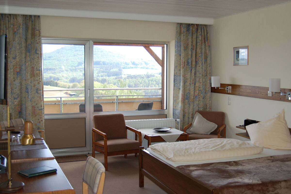 Ein Hotelzimmer mit einem Bett, zwei Stühlen, einem Tisch, einem Schreibtisch und einem Fenster mit Blick auf einen Wald und Berge.