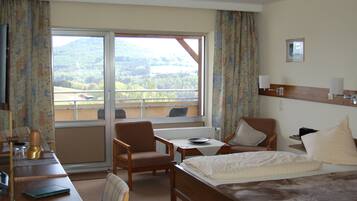 Habitación de hotel con cama, dos sillas, una mesa, un escritorio y una ventana con vistas a un bosque y montañas.