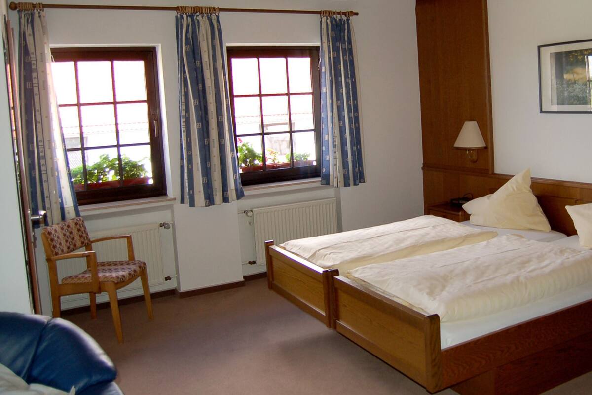 Ein Schlafzimmer mit einem hölzernen Bett, zwei Nachttischen, einem Sessel, einem Fenster mit Vorhängen, einer Heizung und einem gerahmten Bild an der Wand.