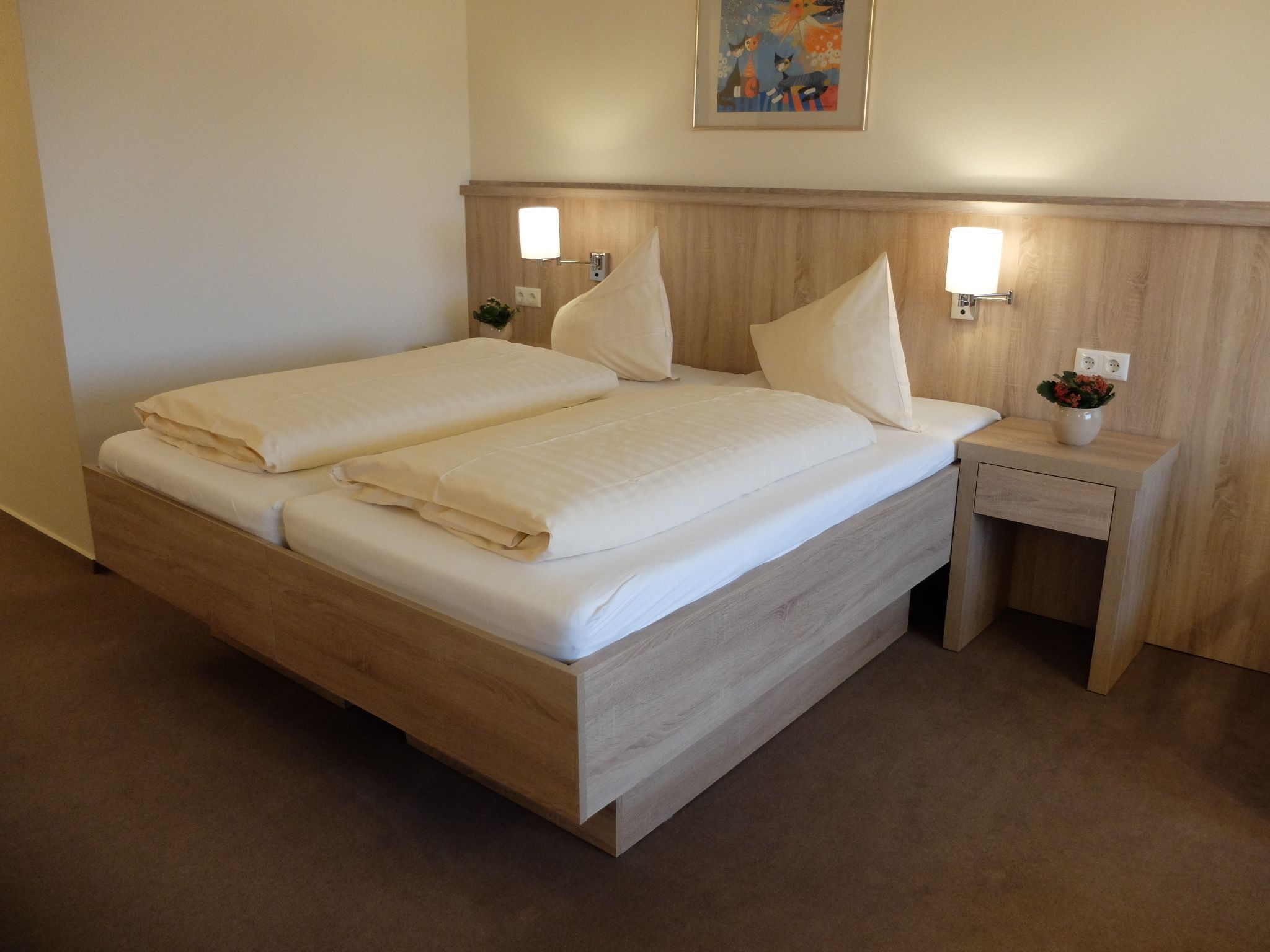 Een opgemaakt tweepersoonsbed met een houten hoofdeinde, twee nachtkastjes met lampen en een kleine vaas met bloemen aan de rechterkant.