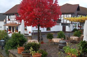 Une cour avec des sièges en plein air, un arbre aux feuilles rouges et un bâtiment doté d’une façade blanche et d’un toit foncé.