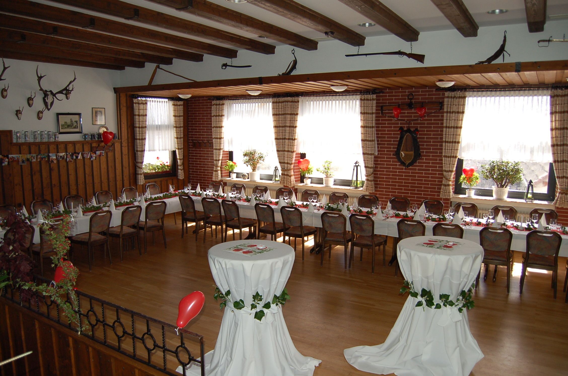Um salão de banquetes com mesas longas preparadas para um evento formal, vigas de madeira no teto e janelas com cortinas.