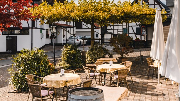 An outdoor café with tables and chairs, a wooden barrel, and a building with a white facade and dark trim in the background. - Landgasthaus Pfahl (Wershofen)
