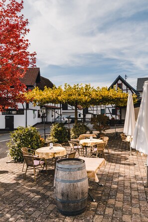 Un café en plein air avec des tables et des chaises, un tonneau en bois, et un bâtiment à l’arrière-plan, doté d’une façade blanche et de bordures sombres.