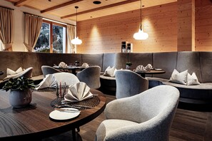A modern dining area with wooden walls, grey upholstered seating, and pendant lighting. - Boutique Hotel Nives (Selva di Val Gardena)