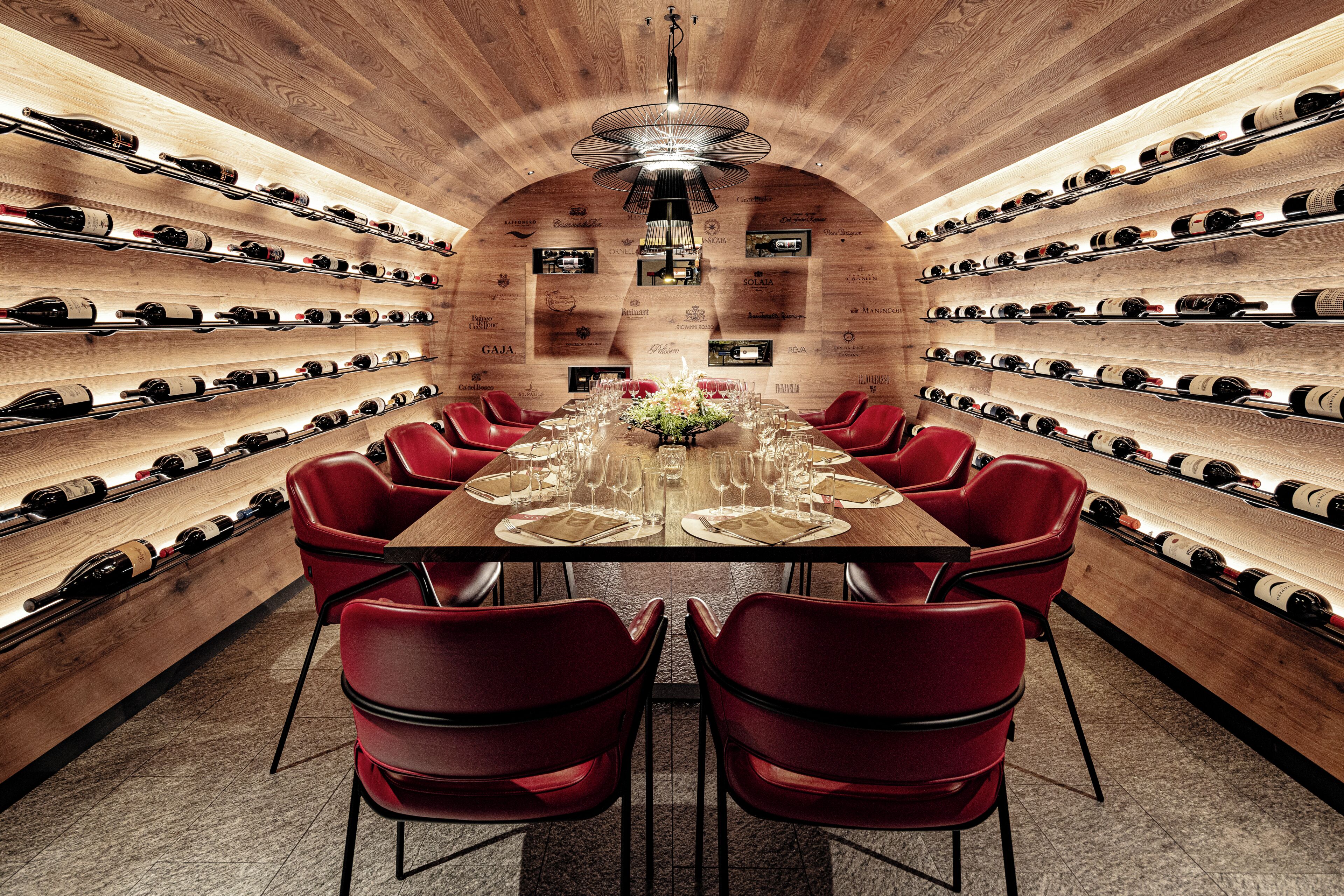 A wine cellar with a long table and red chairs, surrounded by wine racks.