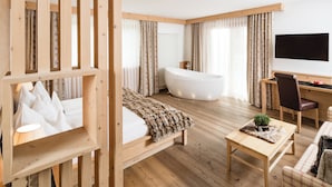 Habitación de hotel moderna con suelo de madera, una bañera grande, una cama con manta blanca, un escritorio con silla y un televisor en la pared.