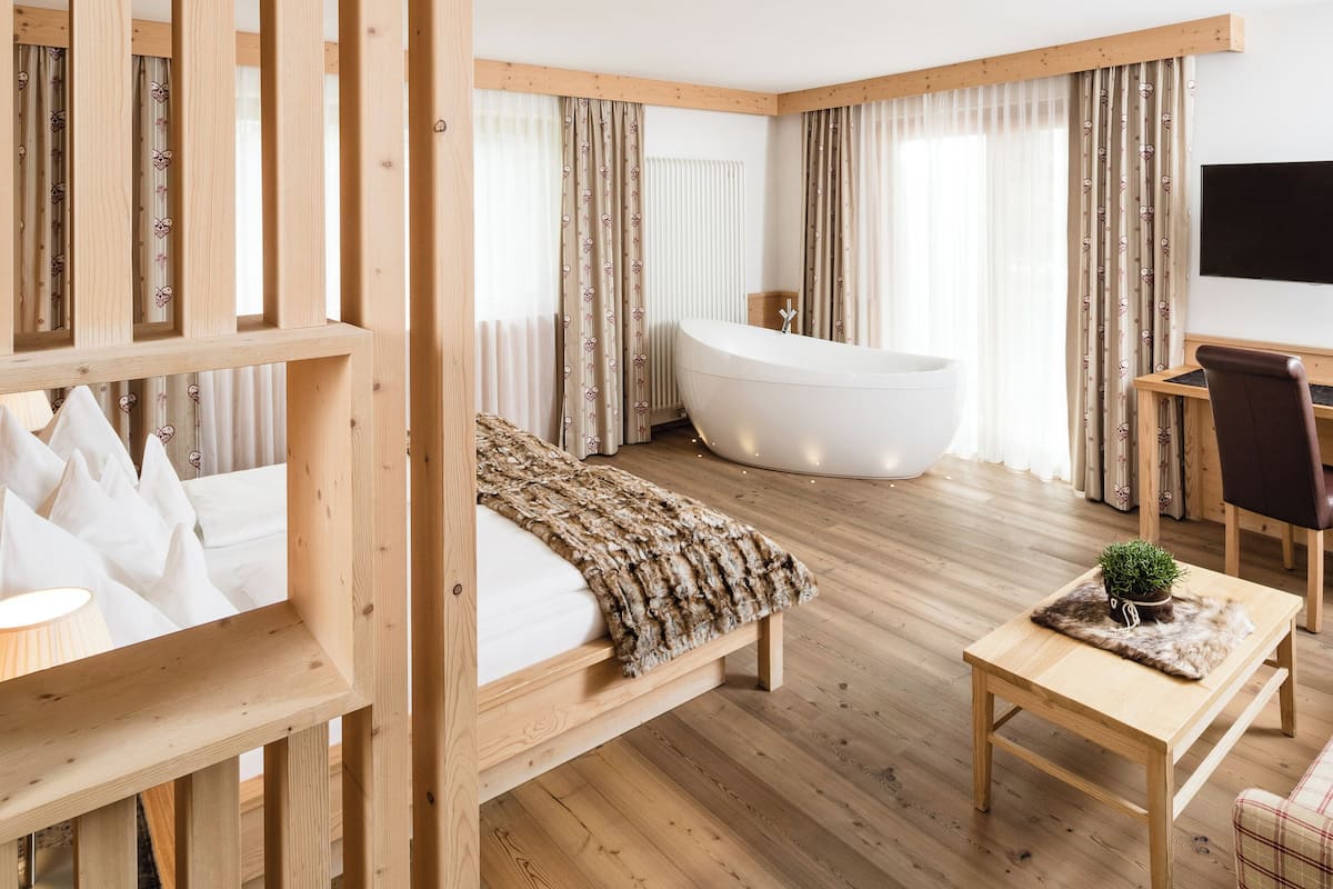A modern hotel room with a wooden floor, a large bathtub, a bed with a white blanket, a desk with a chair, and a television on the wall.