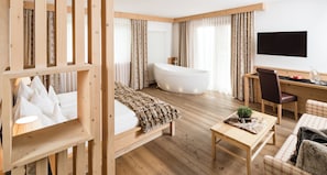 Ein modernes Hotelzimmer mit Holzboden, einer großen Badewanne, einem Bett mit weißer Decke, einem Schreibtisch mit Stuhl und einem an der Wand befestigten Fernseher.