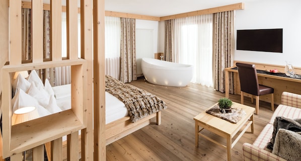 Habitación de hotel moderna con suelo de madera, una bañera grande, una cama con manta blanca, un escritorio con silla y un televisor en la pared.
