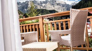 Un balcón con sillas de mimbre y baranda de madera, con vistas a un paisaje montañoso con árboles y cielo despejado.